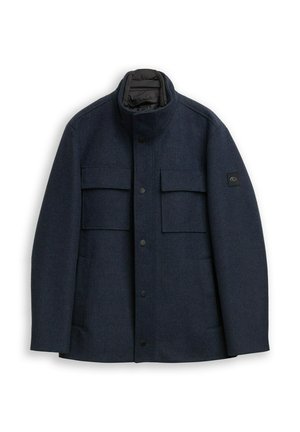 Marineblauer Mantel aus strukturiertem Stoff, mit hohem Kragen, knöpfbarem Vorderteil und zwei Brusttaschen, auf dem Ärmel befindet sich ein Logo-Patch.