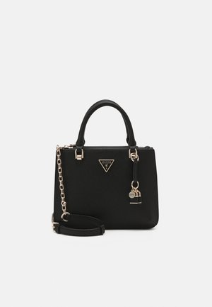 Sac à main noir Guess avec double poignée, bandoulière chaîne dorée et breloque logo doré sur surface lisse et texturée sur fond blanc.