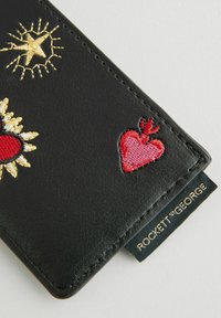 Melnas ādas maku ar izšūtu sarkano sirdi un saules dizainiem. Izceļas ar teksturētu apdari un zelta zīmola etiketi malā.