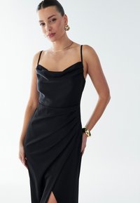 Black satin slip dress with a cowl neckline and thin straps, featuring a draped design, side slit, and smooth texture. Gold jewelry accents.