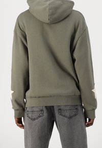 Felpa con cappuccio in cotone verde oliva con un fit rilassato. Presenta accenti bianchi a forma di stella sulle maniche e polsini a costine. Abbinata a jeans in denim grigi.