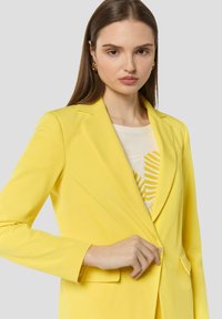 Blazer giallo su misura con una texture liscia, chiusura a un bottone e rever a punta. Indossato sopra una maglietta leggera con stampa di foglie.