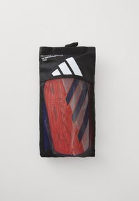Czarna siatkowa torba z widocznymi błyszczącymi ochraniaczami na golenie w kolorach czerwonym i fioletowym. Zawiera logo Adidas oraz tekst określający rodzaj produktu.