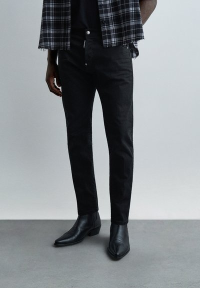 Homme portant un jean noir slim, des bottines noires à bout pointu, et une chemise à carreaux noire et blanche sur un fond gris uni.