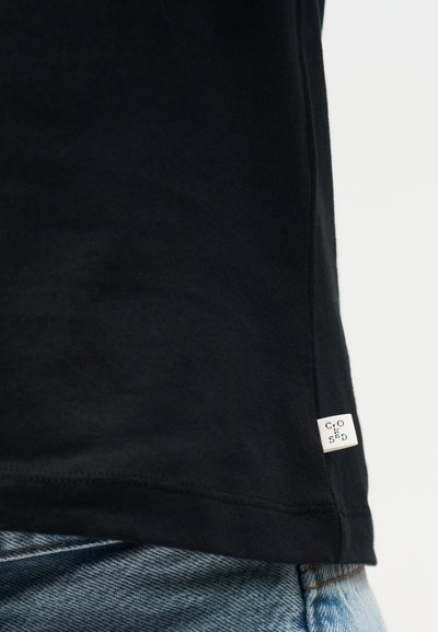 Κοντινό πλάνο στο τελείωμα μαύρου ρούχου με λευκή ετικέτα που γράφει «CLOSED», φορεμένο πάνω από ανοιχτό μπλε τζιν.