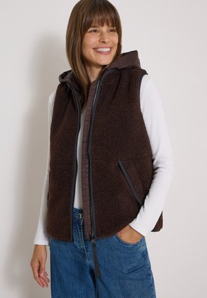 Femme souriante portant une veste polaire marron foncé avec capuche par-dessus une chemise blanche à manches longues et un jean bleu, debout avec une main dans la poche.