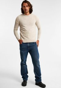 Vaaleanbeige crewneck-paita pitkähihaisilla hihoilla, yhdistettynä sinisiin farkkuihin ja mustiin jalkineisiin, seisoo pystyasennossa neutraalissa taustassa.