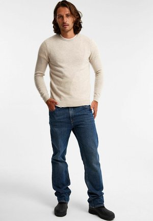 Lichtbeige crewneck sweater met lange mouwen, gecombineerd met blauwe spijkerbroek en zwarte schoenen, rechtop staand tegen een neutrale achtergrond.