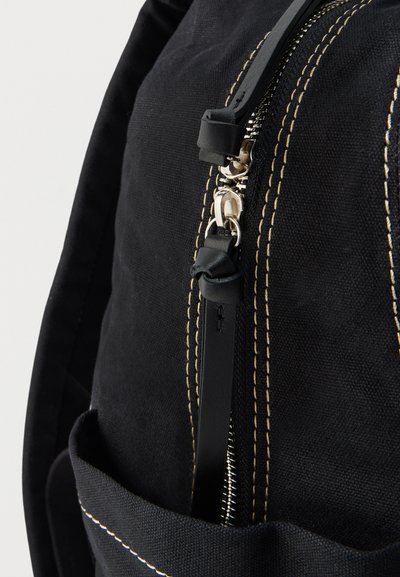 Musta kangaskassi, jossa on beigeä kontrastitikkauksia ja hopeinen vetoketju, jossa musta nahkainen vetolaite solmusomisteella.