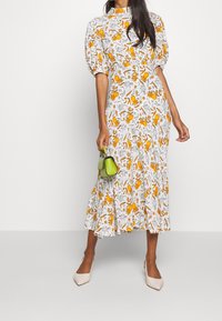 Femme portant une robe midi blanche avec un imprimé floral orange, des manches bouffantes, tenant un petit sac à main vert lime et des chaussures beige à bout pointu.