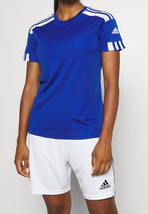 Kobieta ubrana w niebieską sportową koszulkę Adidas z białymi paskami na rękawach i białe szorty Adidas, stoi na jednolitym tle.