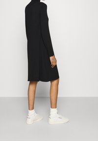 Personne portant une robe noire à manches longues, des chaussettes blanches montantes et des baskets blanches Reebok, debout sur un sol gris uni devant un mur blanc.