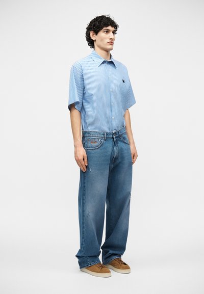 Jeune homme debout, portant une chemise à manches courtes bleu clair à rayures verticales, un jean bleu ample et des baskets marron sur un fond blanc.