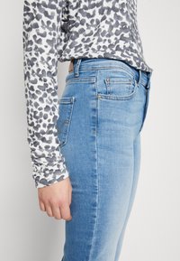 Jean en denim bleu ajusté avec poches, accompagné d'une chemise à manches longues à motif texturé gris et blanc et d'une taille visible.