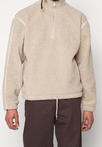 Beige fleece-tröja med hög krage, halv-zip design och sidostreck. Har en dragsko i nederkanten och ett strukturerat, mjukt tyg.