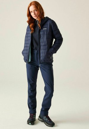 Vrouw met rood haar die een marineblauw gewatteerde jas met capuchon draagt, donkere broek en zwarte wandelschoenen met roze en turquoise accenten, staand tegen een lichte achtergrond.