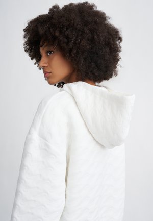 Weißer, gesteppter Hoodie mit einem strukturierten Wellenmuster, ausgestattet mit einer Kapuze und weichem, plüschigem Material. Das Design ist oversized mit überschnittenen Schultern.