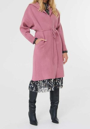 Femme portant un manteau rose ceinturé, une robe à imprimé animal et des bottes noires jusqu’aux genoux, debout devant un fond blanc uni.