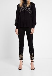 Mujer vestida con una blusa negra de manga larga con un diseño de cuentas en el pecho, pantalones skinny negros con bordado dorado cerca de los tobillos, y tacones negros con tiras.