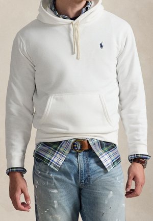 Homme portant un sweat à capuche blanc par-dessus une chemise à carreaux verte et bleue, avec un jean bleu clair éclaboussé de peinture et une ceinture marron avec une boucle ronde.