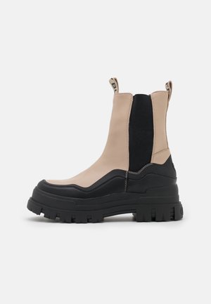 Bottine noire et beige épaisse avec semelle à crampons épaisse, languettes de tirage, panneaux latéraux élastiques et détails cousus sur fond blanc.