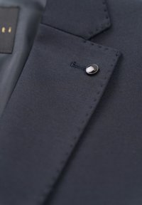 Navy suit jacket detail featuring a smooth texture, distinctive stitching, and a round black button on the lapel.
