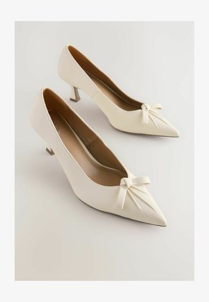Scarpe col tacco alto bianche in pelle, con punta appuntita, un piccolo fiocco decorativo sulla parte anteriore e un tacco slanciato e angolare. Texture liscia.