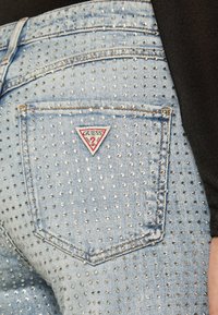 Jeans in denim azzurro con una texture borchiata, con una patch triangolare rossa e bianca sulla tasca posteriore.