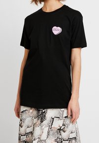 Czarna bawełniana koszulka z krótkim rękawem, z różowym graficznym sercem z napisem "MEMES" na lewej klatce piersiowej. Klasyczny krój.