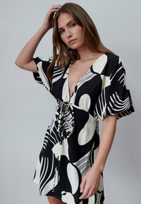 Wickelkleid mit kurzen Ärmeln in Schwarz und Weiß, mit tiefem V-Ausschnitt, Kordelzug an der Taille und abstrakten Blattmustern auf einem strukturierten Stoff.