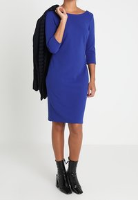 Robe ajustée bleu royal à manches trois-quarts, associée à une veste noire portée sur l'épaule. Bottines noires à talons.