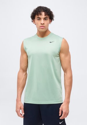 Jeune homme aux cheveux bouclés portant un t-shirt sans manches vert clair Nike et un short foncé, debout devant un fond uni.