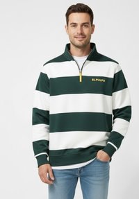 Hombre vistiendo un sudadera de media cremallera de rayas anchas en verde y blanco con el texto "EL PULPO", combinada con jeans de color azul claro, de pie frente a un fondo blanco.