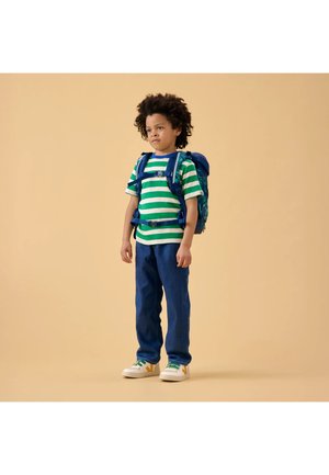 Child with curly hair wearing a green and white striped shirt, blue pants, white sneakers, and a blue backpack standing against a beige background.