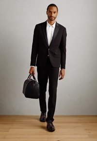 Mann i svart dress og hvit skjorte som går på tregulv, bærende på en svart skinnveske i høyre hånd mot en enkel grå vegg.