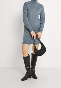 Grå stickad polokragsklänning med långa ärmar, svarta knähöga blanka stövlar och en liten svart handväska med en böjd design.