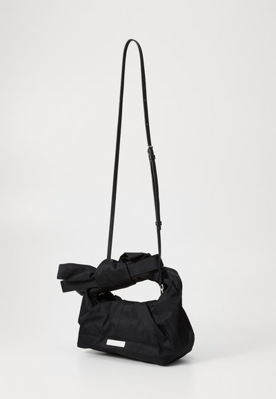 Bolsa de nailon negra con detalle anudado y correa de hombro ajustable. Presenta un cuerpo rectangular y una placa de metal para la etiqueta de la marca.