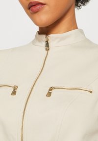 Beige jacket with a high neck, featuring gold zippers on two chest pockets and front. Smooth texture and tailored fit.