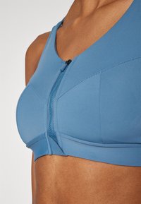 Μπλε αθλητικό σουτιέν με κλείσιμο μπροστά με φερμουάρ, φτιαγμένο από λείο ύφασμα. Διαθέτει ραφές και δομημένο σχέδιο για στήριξη.