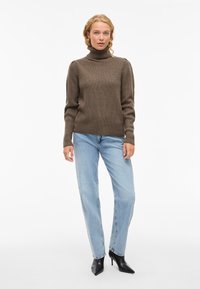 Pulover bej cu guler înalt, cu mâneci lungi, asortat cu blugi drepti de culoare albastru deschis și cizme negre cu toc jos.