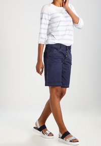 Marineblauwe shorts gecombineerd met een lichtblauw en wit gestreept langemouwen shirt. De outfit bevat casual sandalen met een mix van blauwe en zilveren accenten.