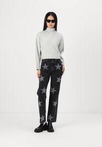 Grå ribbestrikket turtleneck genser, svarte denim jeans med sølvstjernemønster, og svarte sko; modell iført solbriller, stående mot en ensfarget bakgrunn.