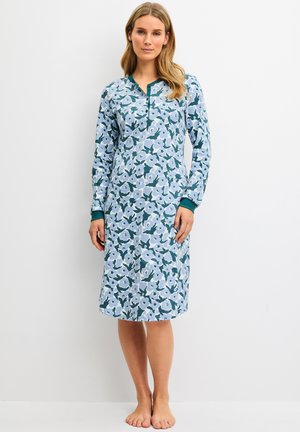 Femme portant une robe à manches longues, longueur genoux avec un motif floral bleu et blanc, debout pieds nus sur un fond blanc uni.