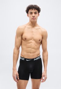 Νεαρός άνδρας με σγουρά μαλλιά φοράει μαύρα εσώρουχα boxer της Nike, στέκεται μπροστά σε λευκό απλό φόντο, κοιτάζοντας κατά μέτωπο με χαλαρή έκφραση.