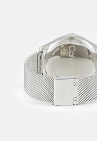 Correa de reloj de malla plateada con un cierre de acero inoxidable pulido. Presenta una caja redonda con "Calvin Klein" grabado en la parte posterior.