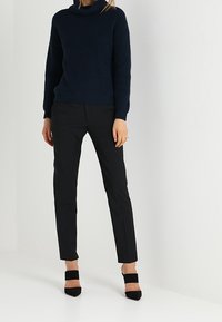 Pull en tricot navy avec col boule, associé à un pantalon noir slim-fit. Le modèle porte des chaussures à talons hauts noires avec des brides de cheville.