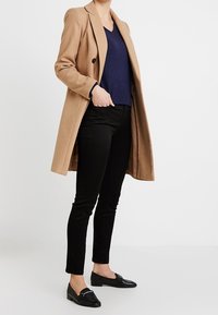Cappotto in misto lana cammello con collo a risvolto, indossato sopra un maglione a V blu navy, pantaloni skinny neri e mocassini neri.