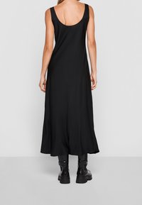 Mujer vistiendo un vestido midi negro sin mangas con un espalda descubierta y botas negras gruesas hasta el tobillo, de pie contra un fondo liso.