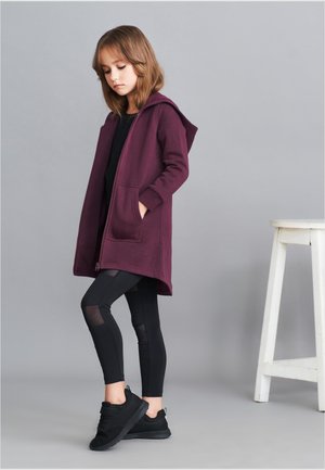 Maroon hoodie med framfickor, ihopparad med svarta leggings med nätdetaljer. Modellen bär svarta sneakers och står bredvid en vit pall.