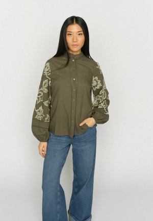 Mujer con cabello largo y negro que lleva una blusa abotonada de color verde oliva con bordados florales blancos en las mangas y vaqueros de pierna ancha azules.